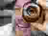 A smiling person holds a glazed chocolate donut up to one eye, peeing through