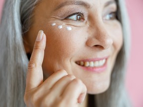Beautiful grey haired Asian lady applies antiaging cream under eye on pink background in studio closeup. Mature beauty lifestyle